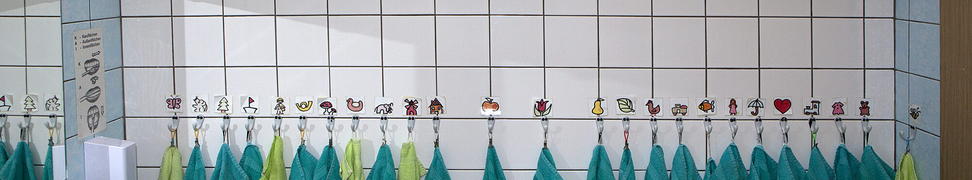 Kindergartenbad: Viele Zahnputzbecher und Handtücher mit Symbolen zur Erkennung, links ein Waschbecken. Oben auf dem Regal ein Demo-Gebiss