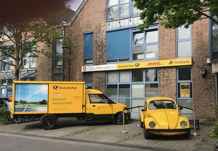 Postagentur in Merzenich - vor dem Gebäude steht rechts ein alter gelber VW Käfer, links ein modernes Post-E-Fahrzeug